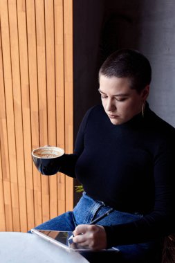 Kısa saçlı ve burun piercingli genç bir kız bir kafede oturuyor ve tablette çalışıyor. Serbest çalışma ve uzak çalışma kavramı..