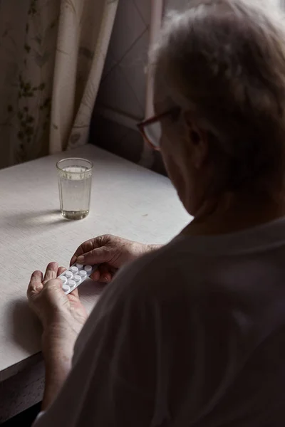 Sad old woman taking pills, health problems in old age, expensive medications. An elderly woman\'s hands unpacking several pills for taking medication. Grandma takes tablet and drinks a glass of water