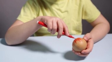Küçük bir çocuk masaya mavi bir arka plan üzerine paskalya yumurtası çiziyor. Küçük çocuk evde boyalı Paskalya yumurtalarını boyuyor. Çocukların Paskalya yaratıcılığı. Paskalya yumurtalarını bir çocuk süslüyor..