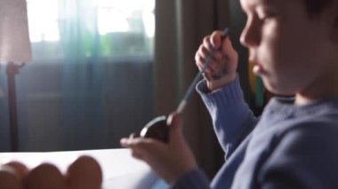 Küçük bir çocuk masaya mavi bir arka plan üzerine paskalya yumurtası çiziyor. Küçük çocuk evde boyalı Paskalya yumurtalarını boyuyor. Çocukların Paskalya yaratıcılığı. Paskalya yumurtalarını bir çocuk süslüyor..