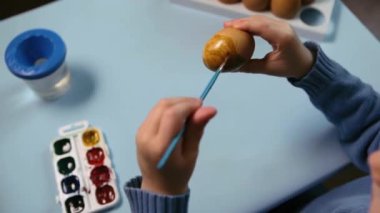 Küçük bir çocuk masaya mavi bir arka plan üzerine paskalya yumurtası çiziyor. Küçük çocuk evde boyalı Paskalya yumurtalarını boyuyor. Çocukların Paskalya yaratıcılığı. Paskalya yumurtalarını bir çocuk süslüyor..