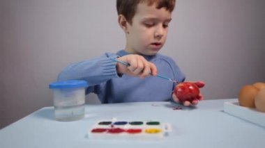 Küçük bir çocuk masaya mavi bir arka plan üzerine paskalya yumurtası çiziyor. Küçük çocuk evde boyalı Paskalya yumurtalarını boyuyor. Çocukların Paskalya yaratıcılığı. Paskalya yumurtalarını bir çocuk süslüyor..