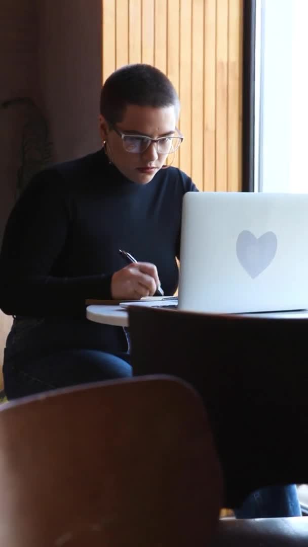 眼鏡をかけた若い女の子が 短い髪でカフェに座ってノートパソコンを見ている 少女は本を見てノートにメモを取る 距離学習の概念 — ストック動画