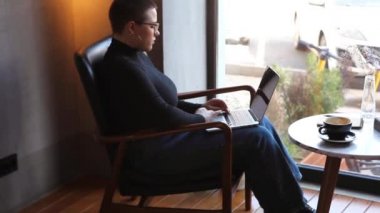 Gözlüklü, kısa saçlı ve burun piercingli genç bir kız bir kafede oturuyor ve bir dizüstü bilgisayar üzerinde çalışıyor. Serbest çalışma ve uzak çalışma veya eğitim kavramı.