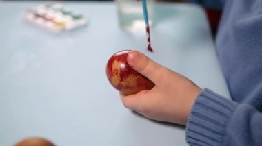 Küçük bir çocuk masaya mavi bir arka plan üzerine paskalya yumurtası çiziyor. Küçük çocuk evde boyalı Paskalya yumurtalarını boyuyor. Çocukların Paskalya yaratıcılığı. Paskalya yumurtalarını bir çocuk süslüyor..