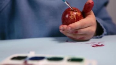 Küçük bir çocuk masaya mavi bir arka plan üzerine paskalya yumurtası çiziyor. Küçük çocuk evde boyalı Paskalya yumurtalarını boyuyor. Çocukların Paskalya yaratıcılığı. Paskalya yumurtalarını bir çocuk süslüyor..