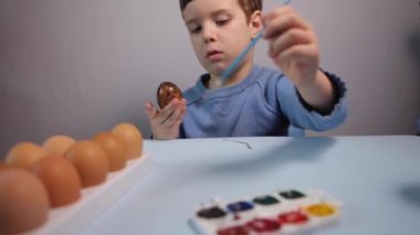Küçük bir çocuk masaya mavi bir arka plan üzerine paskalya yumurtası çiziyor. Küçük çocuk evde boyalı Paskalya yumurtalarını boyuyor. Çocukların Paskalya yaratıcılığı. Paskalya yumurtalarını bir çocuk süslüyor..