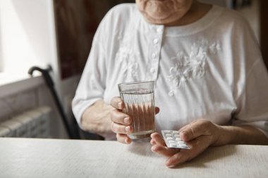 Üzgün yaşlı kadın haplar alıyor, yaşlılıkta sağlık sorunları, pahalı ilaçlar. Yaşlı bir kadının elleri ilaç almak için birkaç hap paketlerini açıyor. Büyükanne tablet alır ve bir bardak su içer.