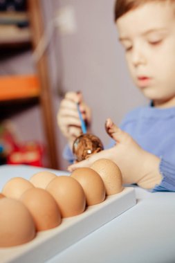 Küçük bir çocuk masaya mavi bir arka plan üzerine paskalya yumurtası çiziyor. Küçük çocuk evde boyalı Paskalya yumurtalarını boyuyor. Çocukların Paskalya yaratıcılığı. Paskalya yumurtalarını bir çocuk süslüyor..