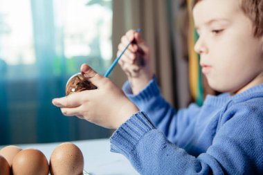 Küçük bir çocuk masaya mavi bir arka plan üzerine paskalya yumurtası çiziyor. Küçük çocuk evde boyalı Paskalya yumurtalarını boyuyor. Çocukların Paskalya yaratıcılığı. Paskalya yumurtalarını bir çocuk süslüyor..