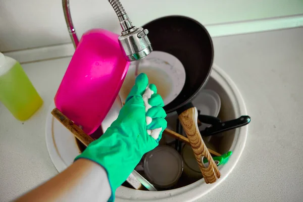 A woman in an apron and rubber gloves washes dirty dishes in the sink in her kitchen. Dirty dishes in a sink for washing up.