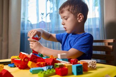 Güzel bir çocuk evde yapı taşlarıyla oynuyor. Şirin, gülümseyen bir çocuk, bir sürü renkli plastik bloğu olan bir inşaatçıyla oynuyor ve bir şehir inşa ediyor. Anaokulu sınıfları.