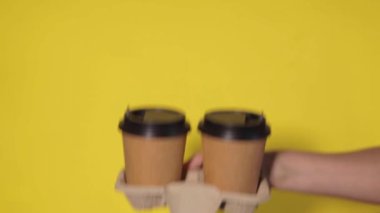 Sarı arka planda yakın plan, kadınların elleri bir stantta kahve ile iki kağıt bardak tutuyor. Seninle kahve ya da çay. Bir fiyatına iki terfi, sarı arka planda kahve teslimatı.