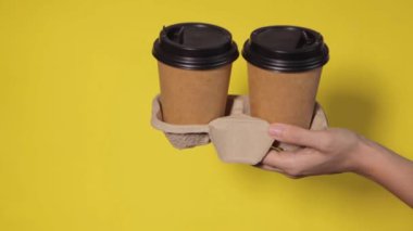 Sarı arka planda yakın plan, kadınların elleri bir stantta kahve ile iki kağıt bardak tutuyor. Seninle kahve ya da çay. Bir fiyatına iki terfi, sarı arka planda kahve teslimatı.