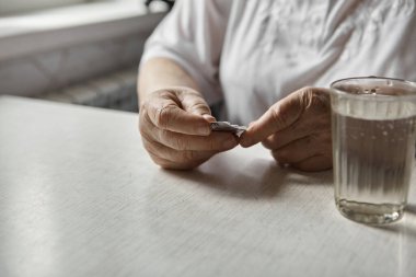 Üzgün yaşlı kadın haplar alıyor, yaşlılıkta sağlık sorunları, pahalı ilaçlar. Yaşlı bir kadının elleri ilaç almak için birkaç hap paketlerini açıyor. Büyükanne tablet alır ve bir bardak su içer.