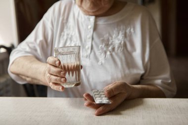 Üzgün yaşlı kadın haplar alıyor, yaşlılıkta sağlık sorunları, pahalı ilaçlar. Yaşlı bir kadının elleri ilaç almak için birkaç hap paketlerini açıyor. Büyükanne tablet alır ve bir bardak su içer.