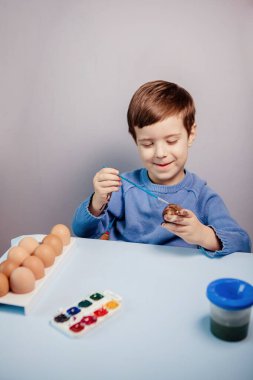 Küçük bir çocuk masaya mavi bir arka plan üzerine paskalya yumurtası çiziyor. Küçük çocuk evde boyalı Paskalya yumurtalarını boyuyor. Çocukların Paskalya yaratıcılığı. Paskalya yumurtalarını bir çocuk süslüyor..