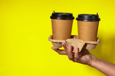 Sarı arka planda yakın plan, kadınların elleri bir stantta kahve ile iki kağıt bardak tutuyor. Seninle kahve ya da çay. Bir fiyatına iki terfi, sarı arka planda kahve teslimatı.