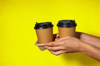 Sarı arka planda yakın plan, kadınların elleri bir stantta kahve ile iki kağıt bardak tutuyor. Seninle kahve ya da çay. Bir fiyatına iki terfi, sarı arka planda kahve teslimatı.