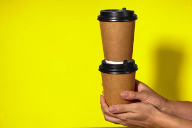 Sarı arka planda yakın plan, kadınların elleri bir stantta kahve ile iki kağıt bardak tutuyor. Seninle kahve ya da çay. Bir fiyatına iki terfi, sarı arka planda kahve teslimatı.