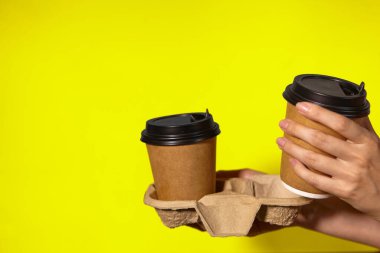 Sarı arka planda yakın plan, kadınların elleri bir stantta kahve ile iki kağıt bardak tutuyor. Seninle kahve ya da çay. Bir fiyatına iki terfi, sarı arka planda kahve teslimatı.