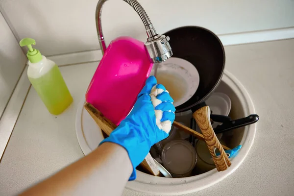 A woman in an apron and rubber gloves washes dirty dishes in the sink in her kitchen. Dirty dishes in a sink for washing up.