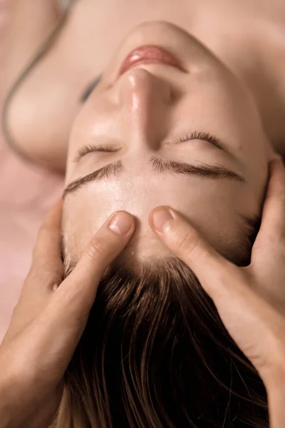 Young beautiful woman is getting a relaxing head and face massage in wellness center. Masseur doing massage the head of an young beautiful girl woman in the spa salon close up macro