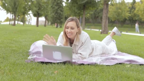 女性は公園のノートパソコンを使って横になっている 笑顔の若い学生の女性は草の上に寝そべってノートパソコンを使用しています 公園で教えるという概念 高品質のフルHd映像 — ストック動画
