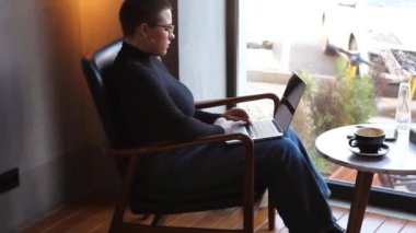 Eğitim ve uzaktan çalışma kavramı. Gözlüklü, kısa saçlı ve burun piercingli genç bir kadın bir kafede oturuyor ve bir dizüstü bilgisayar üzerinde çalışıyor. Serbest çalışma ve uzak çalışma veya eğitim kavramı.