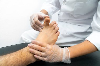 Bir ayak hastalıkları uzmanının sedye üzerinde bir hastayı muayene edişinin yakın çekimi. Doktor eldiven takıyor ve hastanın baş parmağını hareket ettiriyor.