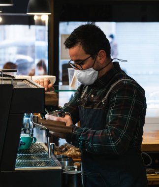 Kafeteryada maske takan beyaz barista kahve makinesinde süt ısıtıyor.