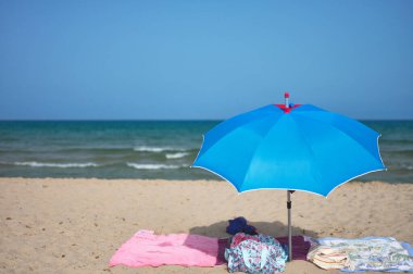 Plajda havluları olan mavi bir şemsiye. Arka planda deniz ve gökyüzü var.