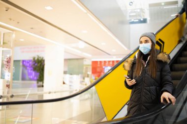Koruyucu maskeli genç bir kadın alışveriş merkezinde merdivenleri kullanıyor.
