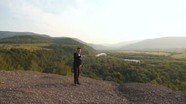 Takım elbiseli genç esmer dağlarda yürüyor. Arka planda güzel bir doğa var. Buluşmayı bekliyor.