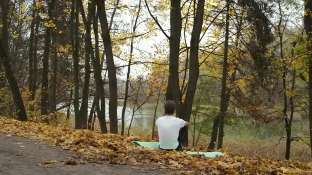 Ένας βάναυσος άντρας κάθεται σε ένα χαλάκι σε ένα φθινοπωρινό πάρκο και περνά από μια όμορφη γέννηση. Διαλογισμός για ανάρρωση. Κατάθλιψη και αυτο-απομόνωση — Αρχείο Βίντεο