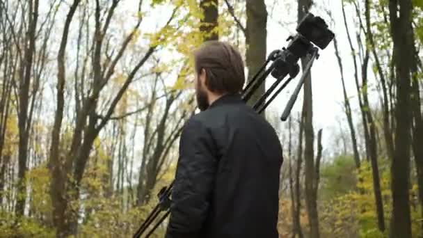 Πορτραίτο ενός βάναυσου φωτογράφου που περπατά μέσα στο φθινοπωρινό δάσος με μια φωτογραφική μηχανή και ένα τρίποδο και φωτογραφίζει τα τοπία του χρυσού φθινοπώρου — Αρχείο Βίντεο