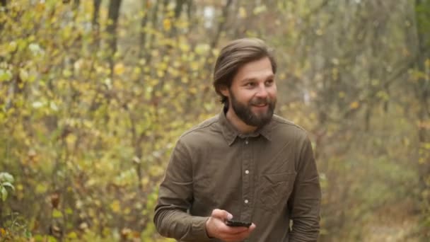 Un uomo brutale sorridente con una camicia marrone cammina nel parco autunnale tenendo in mano un telefono e comunica sui social network. Persona felice — Video Stock