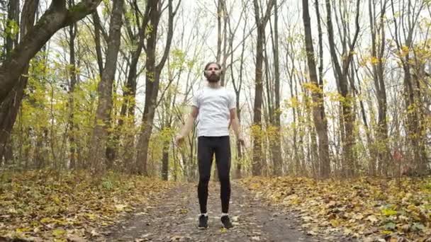 Atleta com fones de ouvido verdes ouve música no parque e aquece antes de correr. Esportes e saúde — Vídeo de Stock