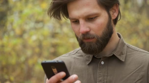 Leende brutal man i brun skjorta går i höstparken med en telefon i händerna och kommunicerar på sociala nätverk. Lycklig person — Stockvideo