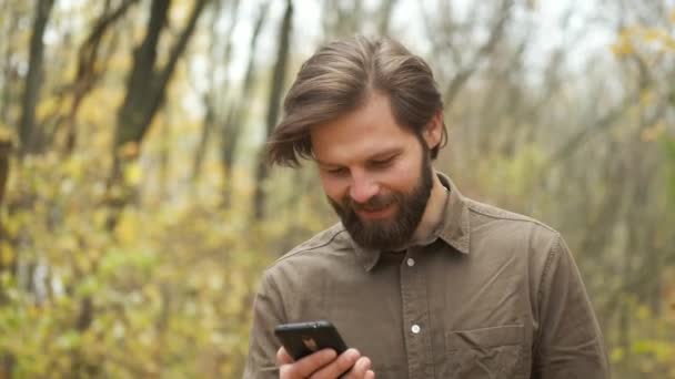 Usmívající se muž v hnědé košili kráčí podzimním parkem, v rukou drží telefon, prohledává internet a objednává si online donášku — Stock video