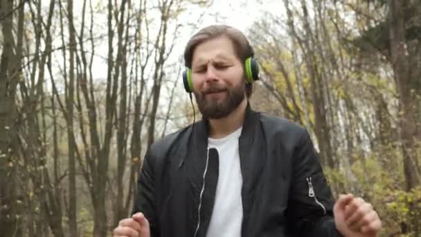 Молодий жорстокий кавказький чоловік емоційно ходить осіннім парком і слухає музику на навушниках. Танці й відпочинок — стокове відео