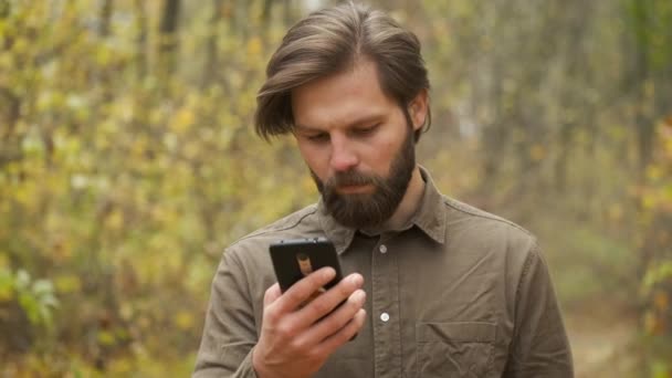 Un uomo brutale sorridente con una camicia marrone cammina nel parco autunnale tenendo in mano un telefono e comunica sui social network. Persona felice — Video Stock