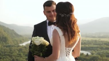 Takım elbiseli damat geline yaklaşır ve çiçekleri verir. Dağların ve nehrin arka planında mutlu bir düğün