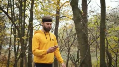 Gülümseyen beyaz bir adam, elinde telefonla sonbahar parkında yürüyor ve sosyal ağlar üzerinden iletişim kuruyor. Mutlu kişi