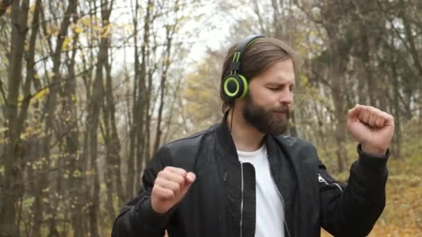Un joven y brutal hombre caucásico está caminando emocionalmente en un parque de otoño y escuchando música con auriculares. Baile y relajación — Vídeo de stock