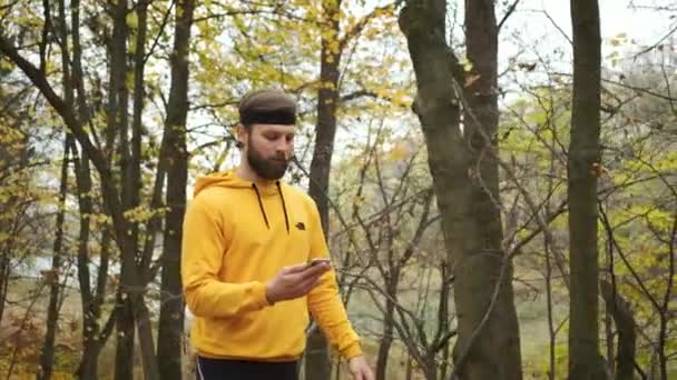 Un hombre caucásico sonriente camina en el parque de otoño, sosteniendo un teléfono en sus manos y se comunica en las redes sociales. Persona feliz — Vídeo de stock