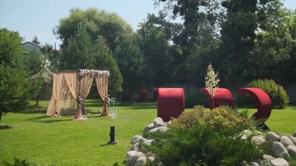 Bröllopsinredning. Vacker bröllopsbåge med färska blommor och röda fåtöljer i trädgården på en sommar solig vacker bröllopsdag — Stockvideo