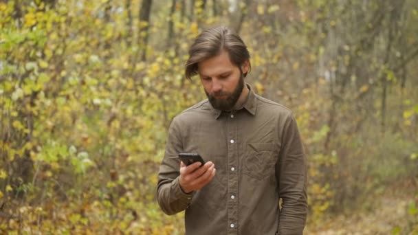 Leende brutal man i brun skjorta går i höstparken med en telefon i händerna och kommunicerar på sociala nätverk. Lycklig person — Stockvideo