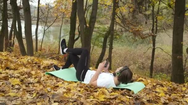 Una morsa caucásica barbuda yace en un parque frío de otoño en una alfombra con una camiseta blanca y escucha emocionalmente música con auriculares verdes. Endurecimiento en el frío — Vídeos de Stock