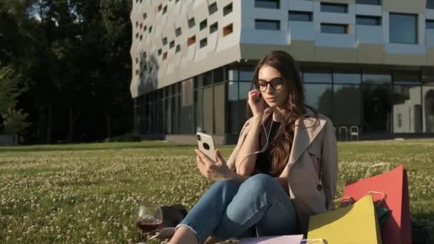 25-jarige mooie brunette met een stijlvolle bril zit op het groene gras in het park op de achtergrond van een modern gebouw en dirigeert een video-uitzending. Werk — Stockvideo